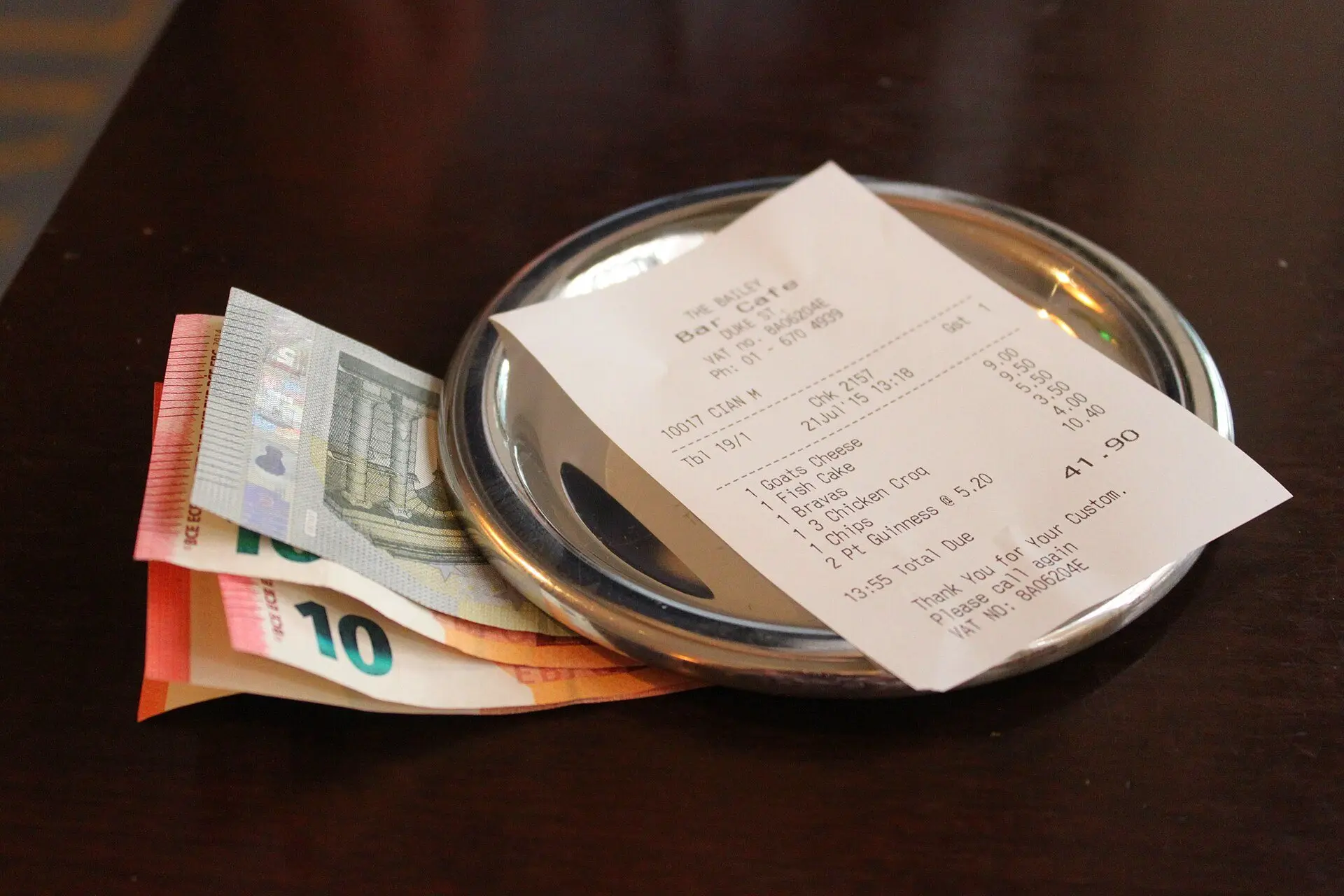 Struk restoran di atas nampan logam kecil, di samping uang kertas euro